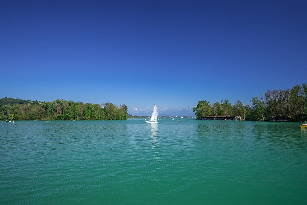 Voilier à voile blanche sur le lac de Sempach entre des rives boisées, sous un ciel bleu